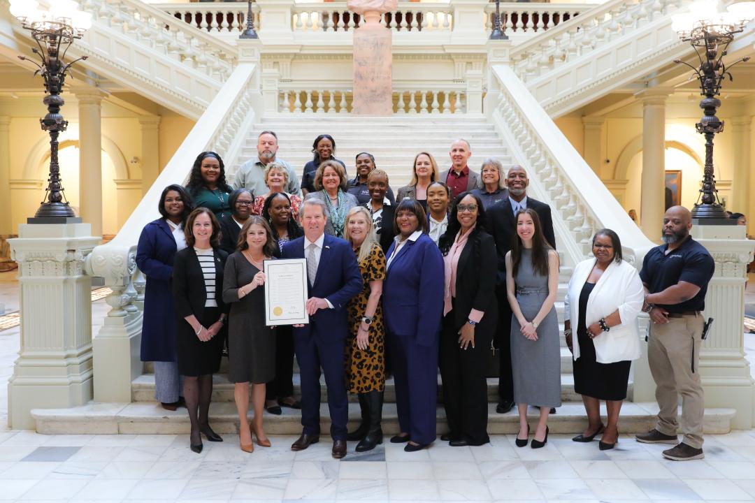 Photo depicts Asset Managers, DOAS Staff including DOAS Commissioner Rebecca Sullivan standing with Governor Brian Kemp and Georgia's First Lady Marty Kemp to celebrate Asset Awareness Month. Governor Kemp and DOAS Commissioner Sullivan stand at the front of the group holding the 2026 Asset Awareness Month Proclamation. Picture is taken in the Georgia Capitol lobby in front of the grand staircase. 