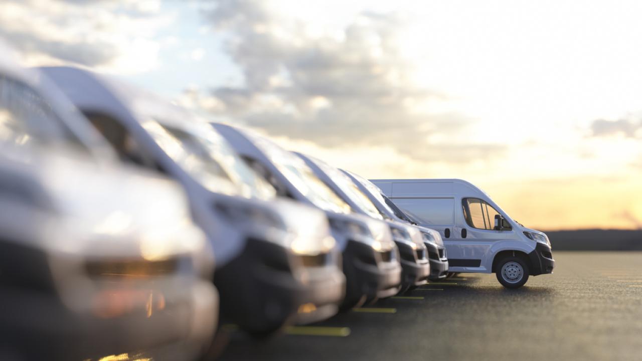 A van slowly drives forward from a line of vans in a sunlit parking lot.