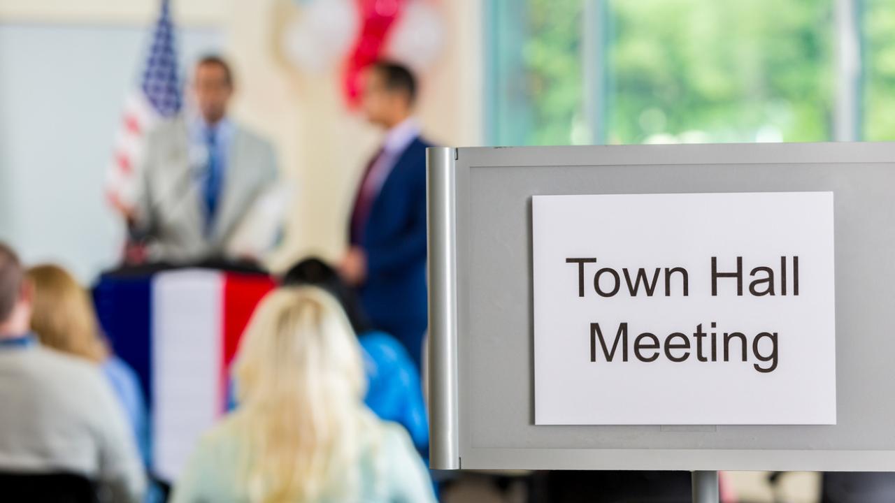 Image of a town hall meeting. In the foreground, a sign reads, &quot;Town Hall Meeting.&quot; The background is blurred, but figures are visible at a podium and in the audience.