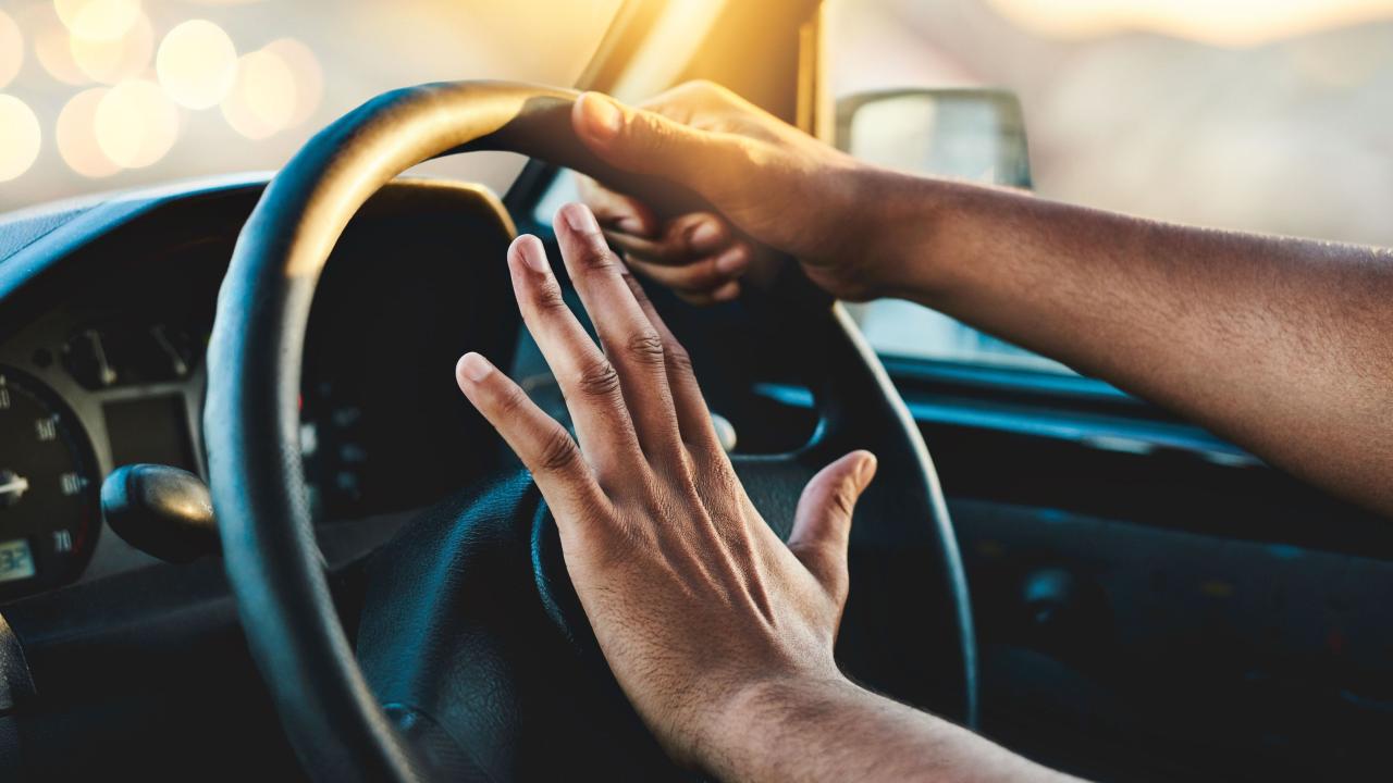 Image of a pair of hands on a steering wheel, on of which is applying the horn.