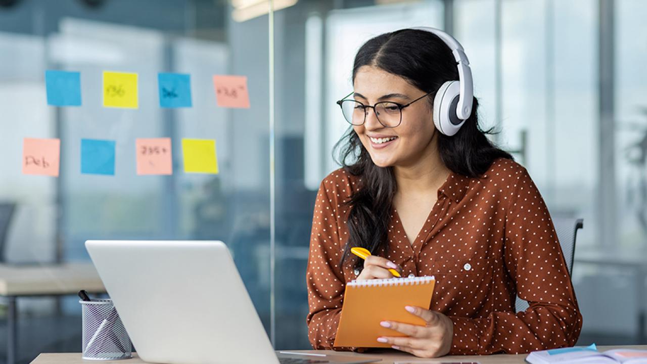 A woman sits at a desk wearing headphones and taking notes while participating in an online learning course.