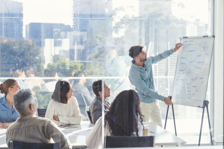 Business people watching a presentation on the whiteboard. A man is writing on the whiteboard with charts and graphs. They are sitting in a board room, there are laptop computers and technology on the table. All are casually dressed. There is a window behind him with city views.