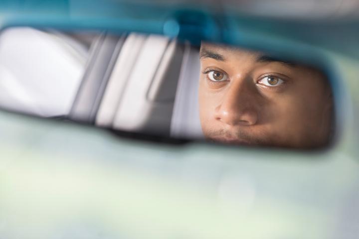 Image of a car&#039;s rear-view mirror, with a man looking into it. 