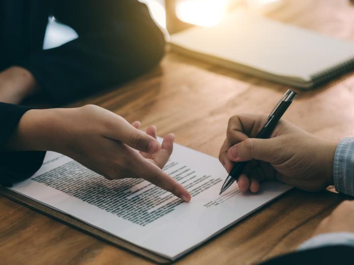 A hand points to the "sign here" line in a contract while another hand holds a pen ready to sign.