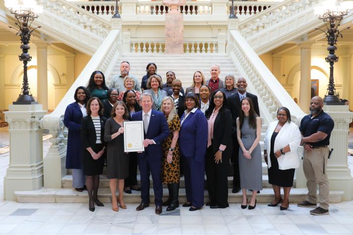 Photo depicts Asset Managers, DOAS Staff including DOAS Commissioner Rebecca Sullivan standing with Governor Brian Kemp and Georgia's First Lady Marty Kemp to celebrate Asset Awareness Month. Governor Kemp and DOAS Commissioner Sullivan stand at the front of the group holding the 2026 Asset Awareness Month Proclamation. Picture is taken in the Georgia Capitol lobby in front of the grand staircase. 