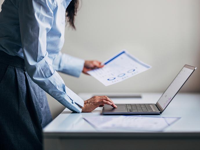A woman stands near a table holding a report while using a laptop computer.